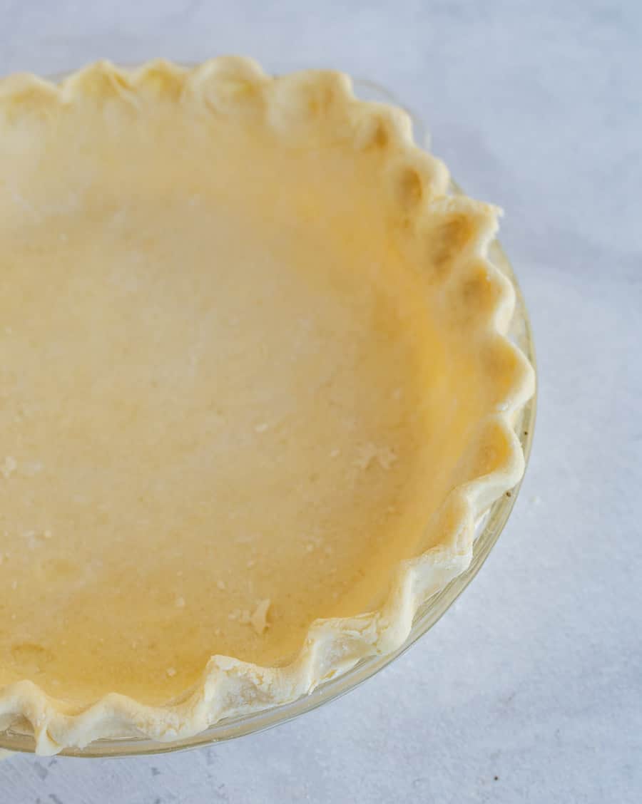 A clear glass pie dish filled with an unbaked pie crust, featuring crimped edges, sits on a light-colored surface—perfect for those learning how to make pie crust from scratch.
