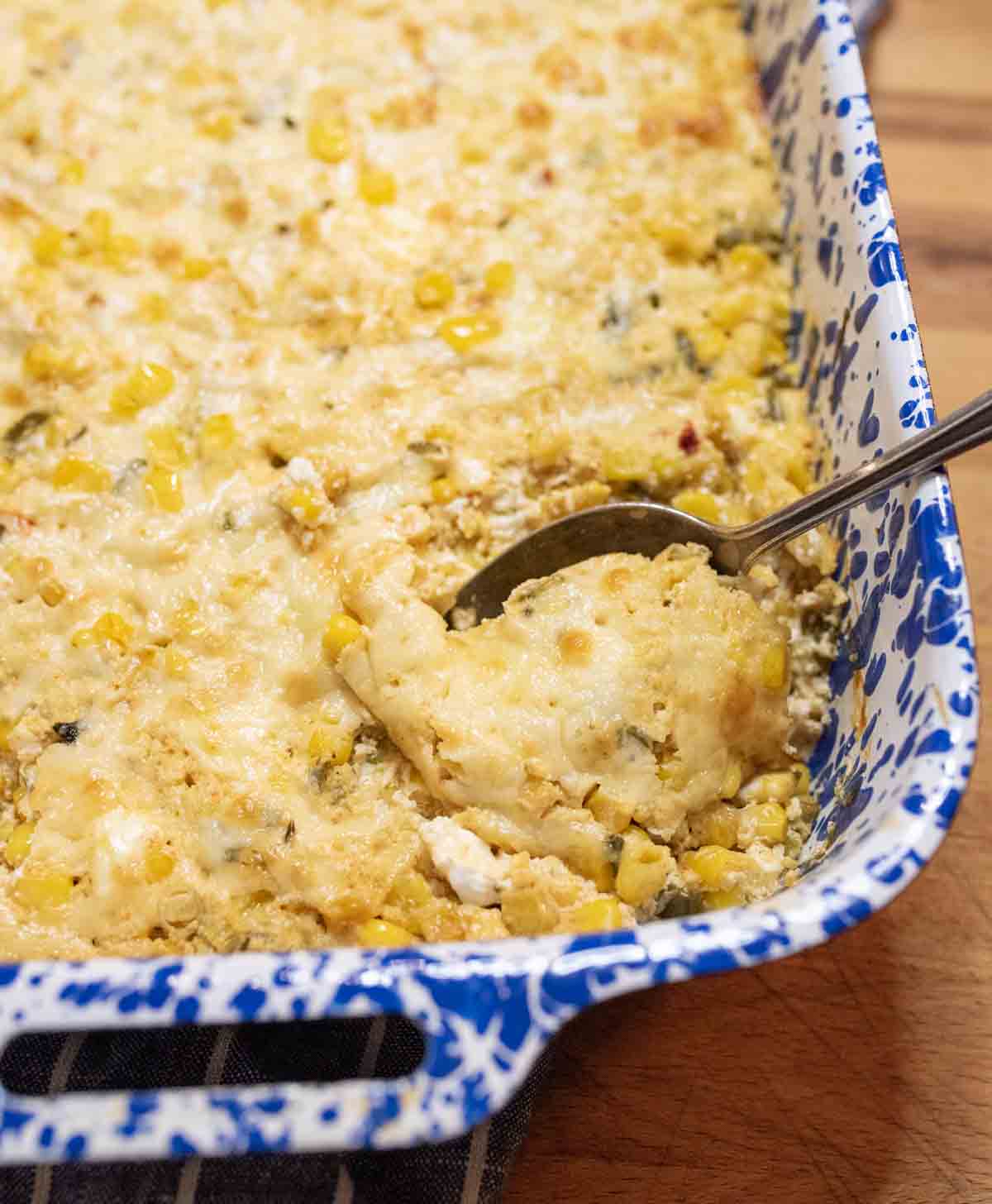 A blue and white baking dish filled with a cheesy corn casserole. A serving spoon is lifting a portion, revealing a creamy, golden-brown mixture with visible corn kernels.