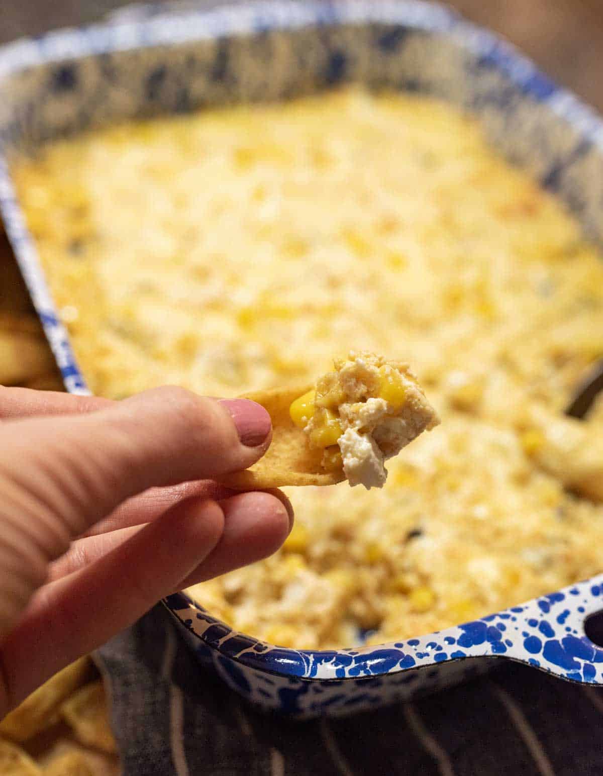 A hand holds a chip topped with creamy, cheesy corn dip in front of a blue and white speckled baking dish filled with more of the dip.