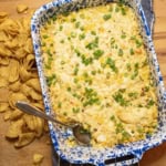A blue and white speckled baking dish filled with creamy corn dip, topped with chopped green onions. A spoon rests in the dish, and corn chips are scattered on the wooden surface alongside.