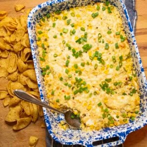 A blue and white speckled baking dish filled with creamy corn dip, topped with chopped green onions. A spoon rests in the dish, and corn chips are scattered on the wooden surface alongside.
