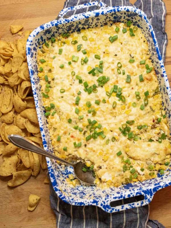 A blue and white speckled baking dish filled with creamy corn dip, topped with chopped green onions. A spoon rests in the dish, and corn chips are scattered on the wooden surface alongside.