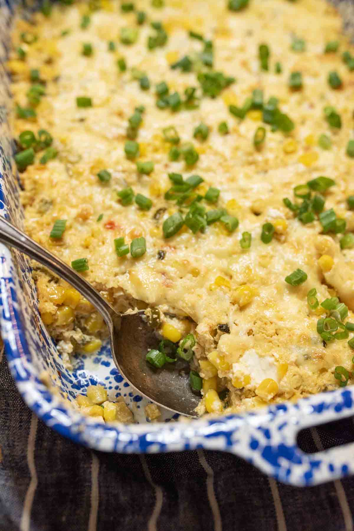 A baked casserole topped with melted cheese, chopped green onions, and corn, partially served with a metal spoon in a blue and white patterned dish.