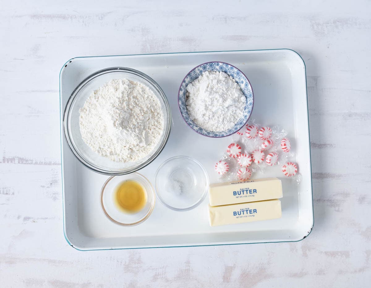 A white tray with ingredients: a bowl of flour, a bowl of powdered sugar, wrapped peppermint candies, two sticks of butter, a small bowl of vanilla extract, and a small dish of salt on a white surface.