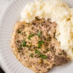 A seasoned, cooked lemon pepper pork chop garnished with fresh herbs sits next to a serving of mashed potatoes on a white plate. A bunch of parsley is partially visible in the background.