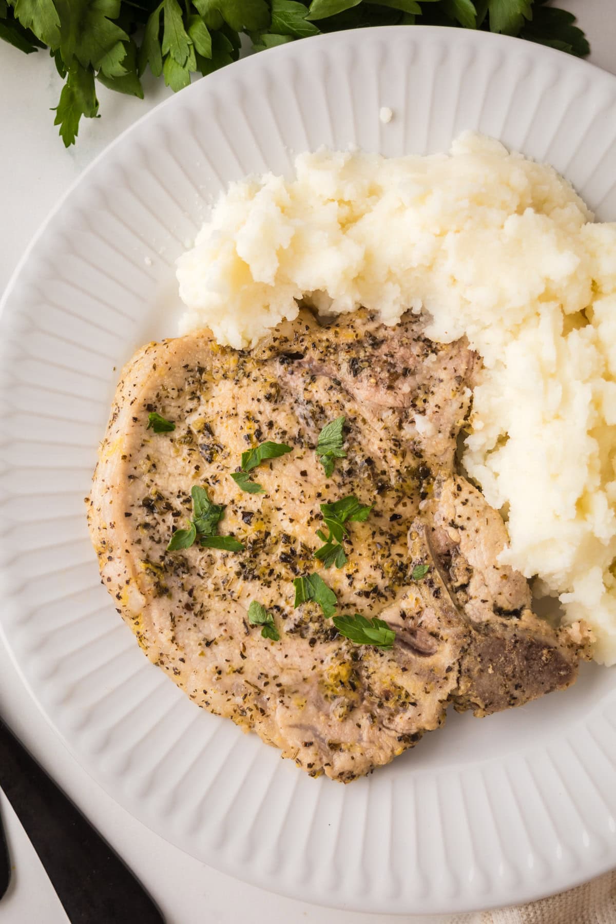 A seasoned, cooked lemon pepper pork chop garnished with fresh herbs sits next to a serving of mashed potatoes on a white plate. A bunch of parsley is partially visible in the background.