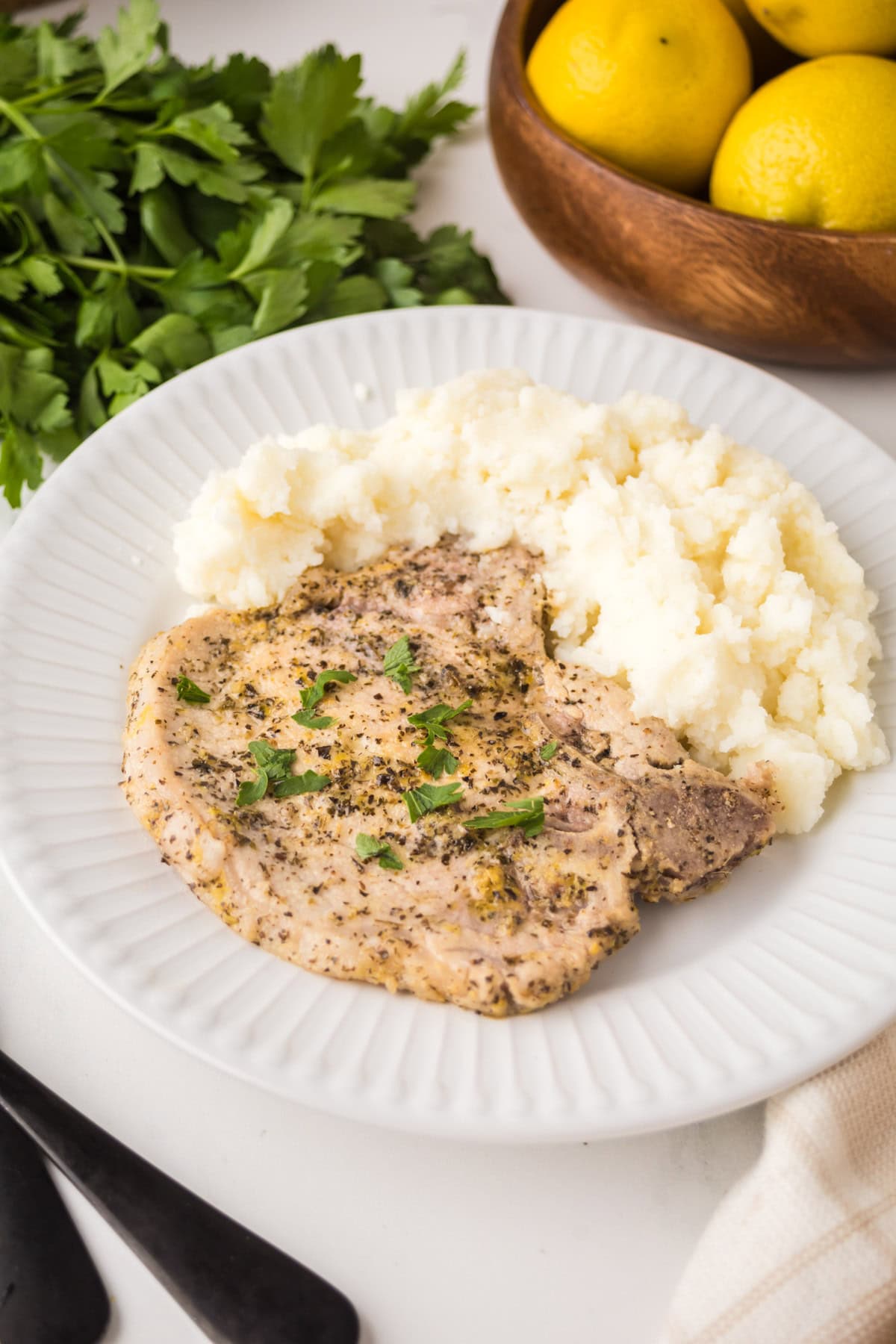 A white plate with a serving of seasoned grilled chicken breast garnished with chopped herbs, next to mashed potatoes. Fresh parsley, a bowl of lemons, and lemon pepper pork chops are in the background.