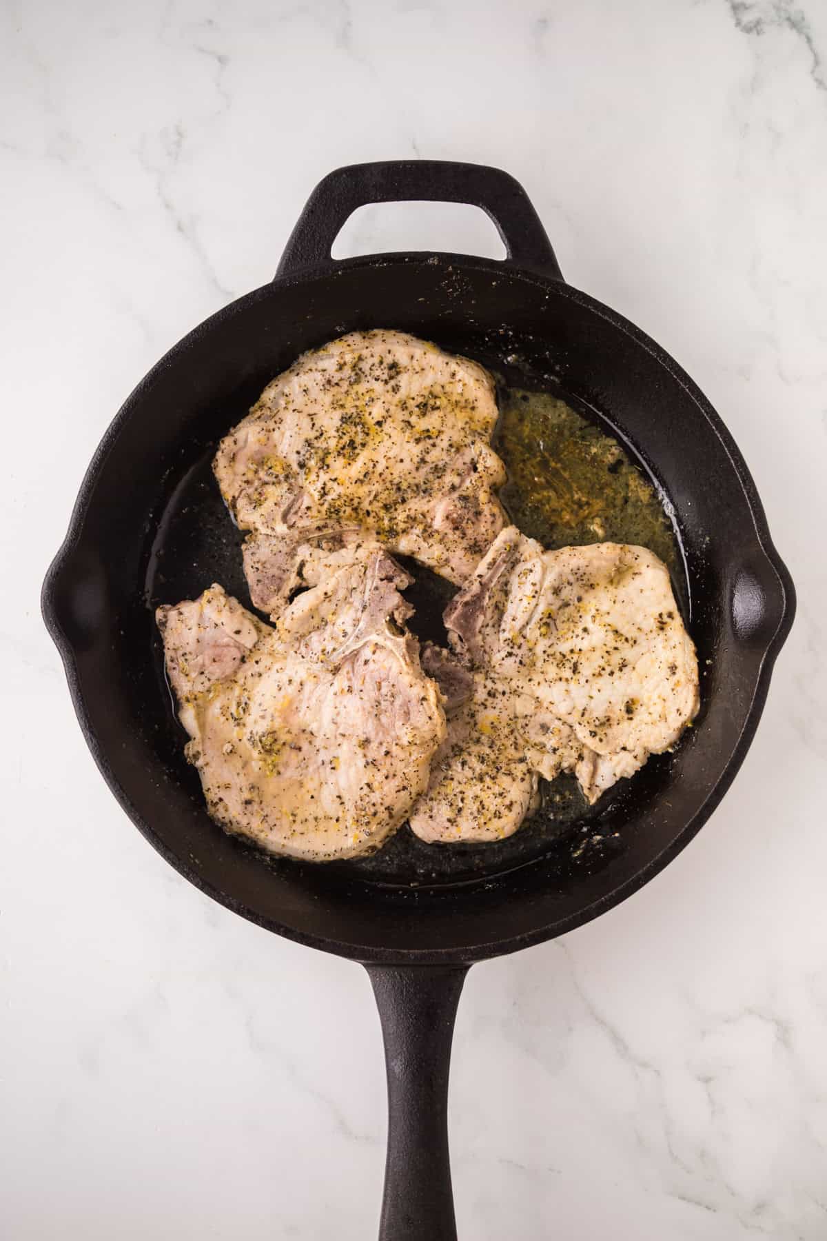 Three seasoned lemon pepper pork chops are cooking in a black cast iron skillet on a white marble countertop.