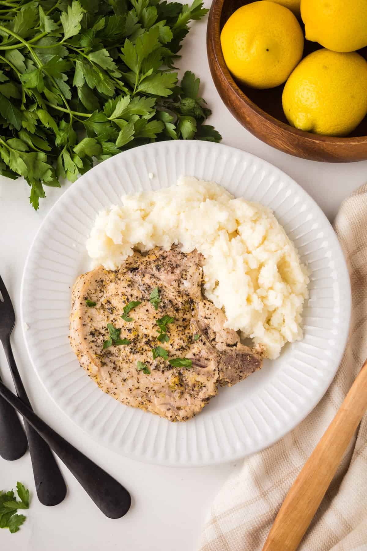 A white plate features lemon pepper pork chops topped with herbs and a serving of mashed potatoes, set on a white table with fresh parsley, lemons in a wooden bowl, black cutlery, and a beige napkin nearby.