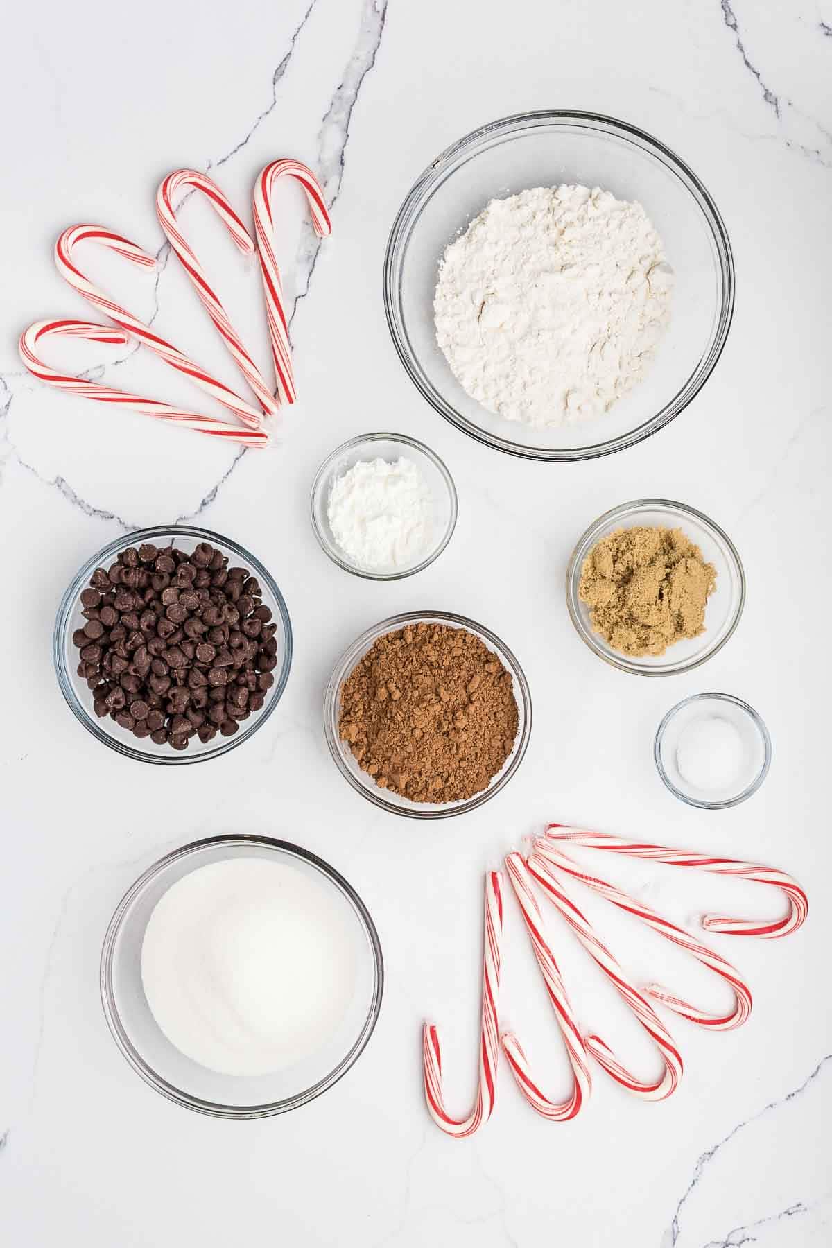Eight candy canes and seven glass bowls containing flour, brown sugar, cocoa powder, chocolate chips, white sugar, salt, and baking powder arranged on a white marble surface.