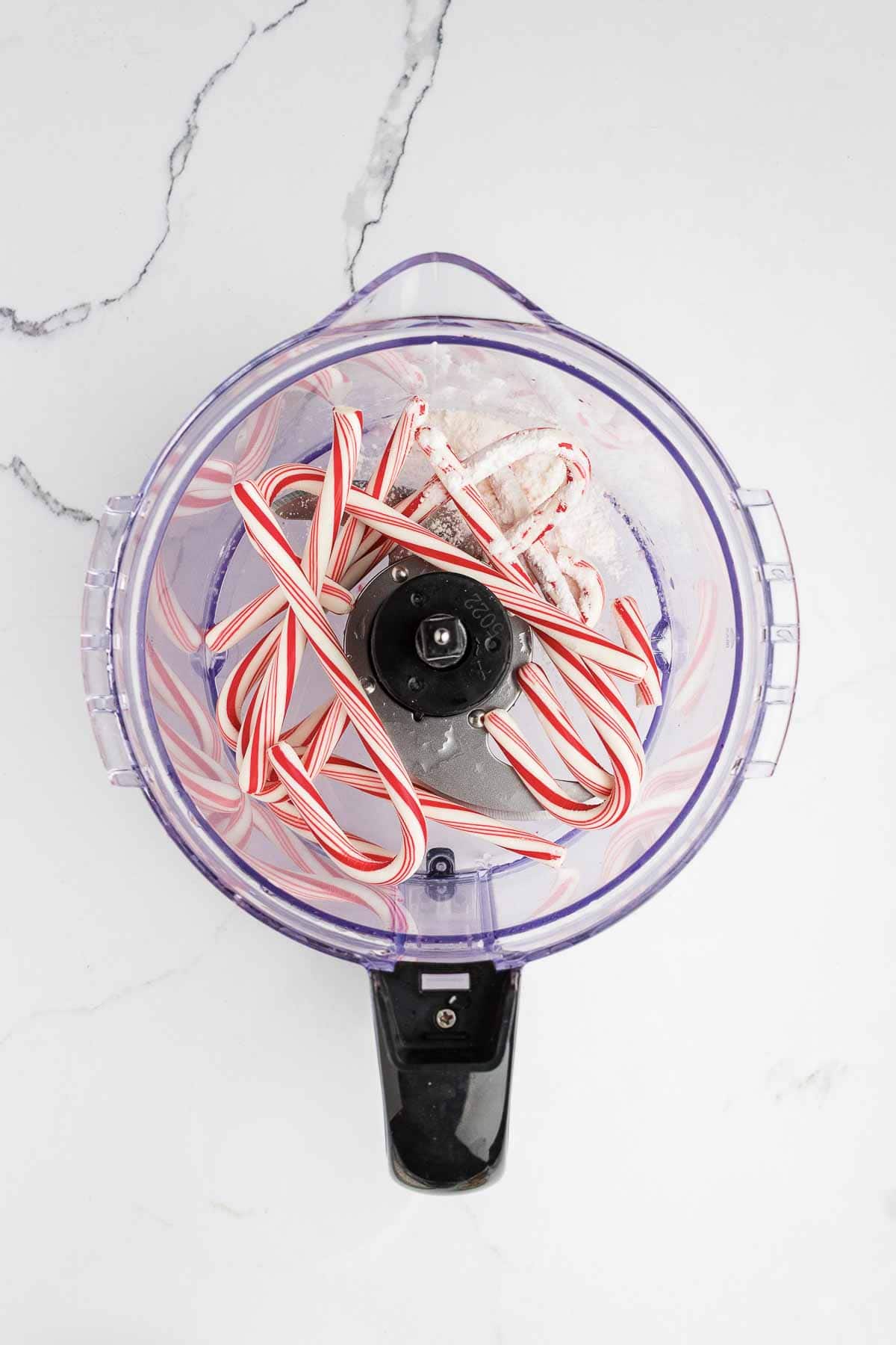A top-down view of a clear food processor bowl containing several whole mini candy canes and white powdered sugar, placed on a white marble surface.