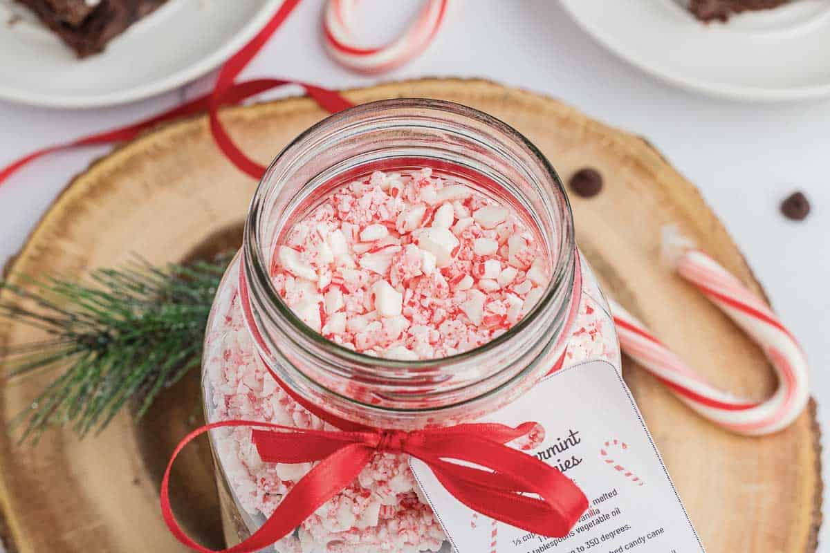 A jar filled with crushed peppermint candies, tied with a red ribbon, sits on a wooden plate with a pine sprig. Candy canes and a plate of brownies are visible in the background.