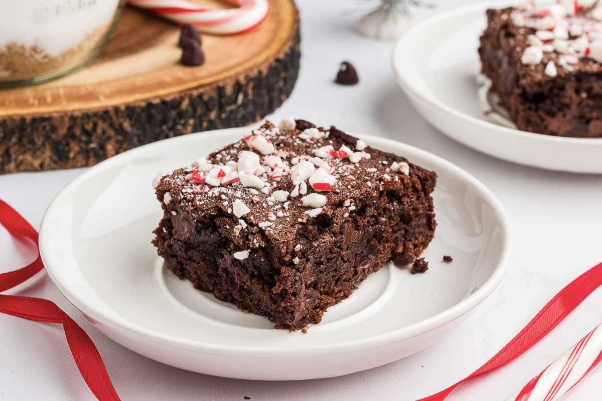 A square piece of chocolate brownie topped with crushed peppermint candy sits on a white plate, with another plate of brownie and a candy cane visible in the background.