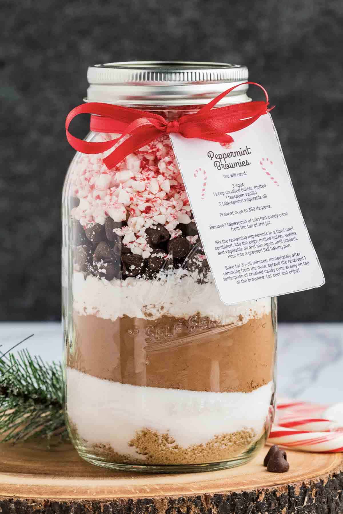 A glass jar filled with layered dry ingredients for peppermint brownies, topped with crushed candy canes and chocolate chips. The jar is tied with a red ribbon and has a recipe card attached. A small pine sprig and candy canes are nearby.