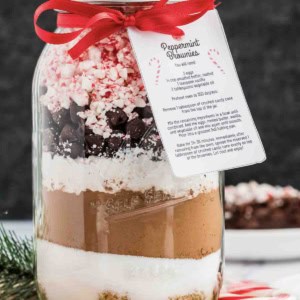 A mason jar filled with layered baking ingredients for peppermint chocolate brownies, topped with crushed peppermint, chocolate chips, and marshmallows, decorated with a red ribbon and a recipe card labeled “Peppermint Brownies.”.