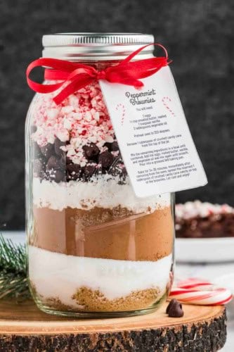 A mason jar filled with layered baking ingredients for peppermint chocolate brownies, topped with crushed peppermint, chocolate chips, and marshmallows, decorated with a red ribbon and a recipe card labeled “Peppermint Brownies.”.