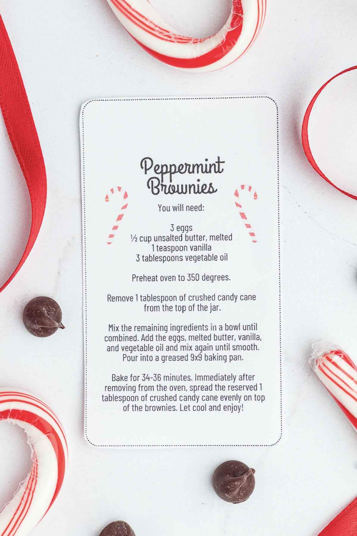A recipe card titled Peppermint Brownies sits on a white surface, surrounded by candy canes, red ribbon, and chocolate chips. The card lists ingredients and instructions for making peppermint brownies.
