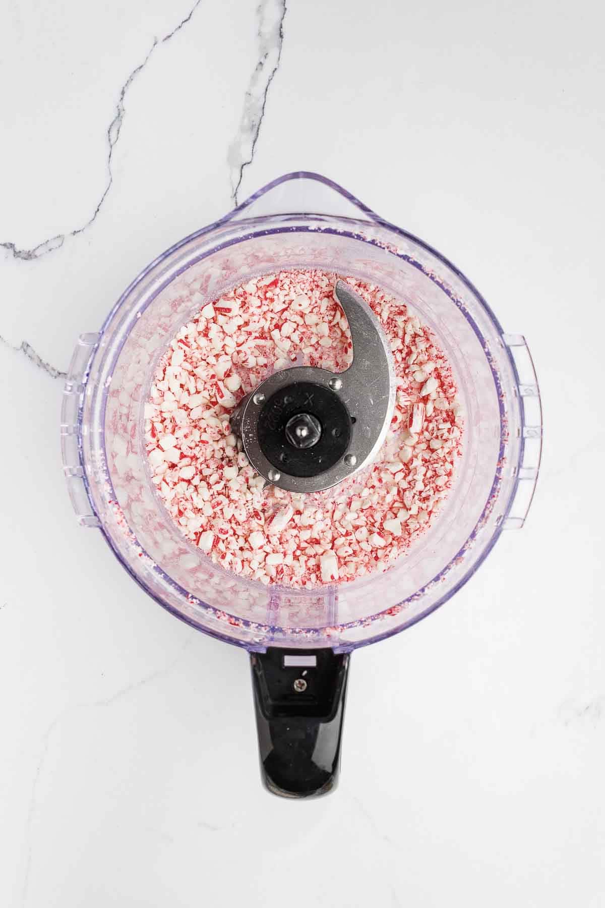 A food processor containing crushed peppermint candies, viewed from above on a white marble surface.