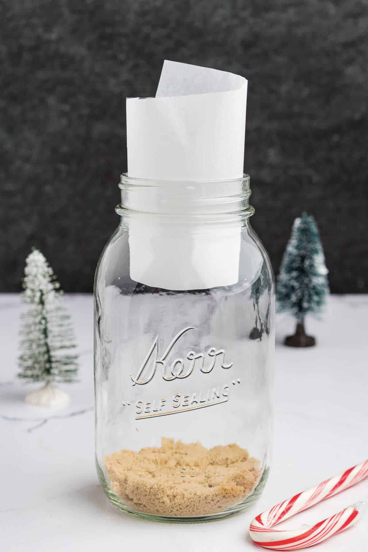 A glass mason jar with brown sugar at the bottom and a rolled piece of white parchment paper inside. There are small decorative pine trees and a candy cane nearby on a white surface.