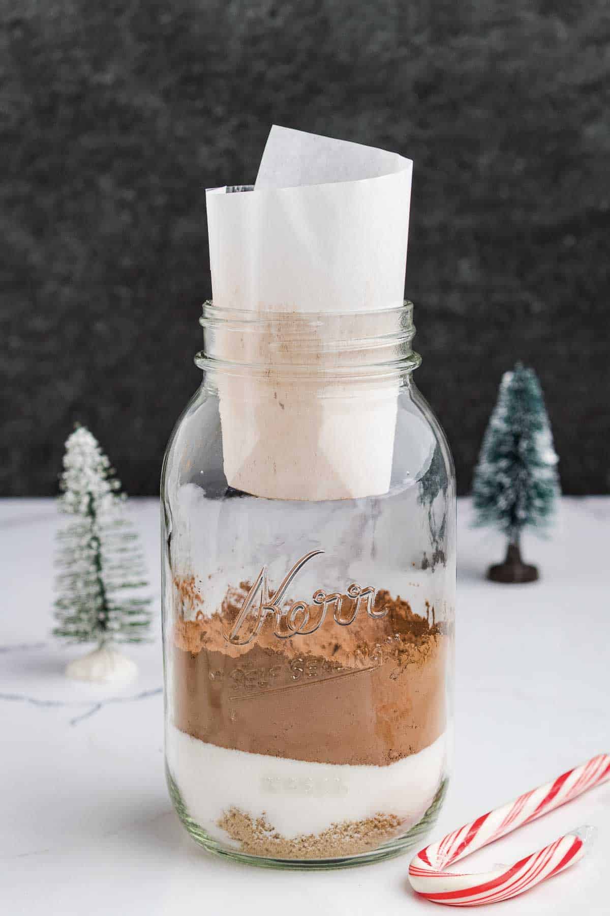 A mason jar filled with layered dry baking ingredients sits on a white surface, with a rolled paper inside the jar. Two mini Christmas trees and a candy cane are nearby, against a dark background.