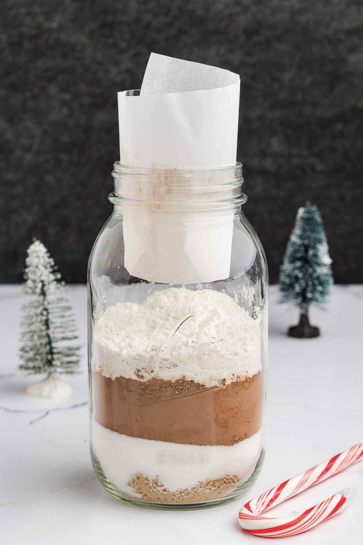A glass jar filled with layered dry ingredients, including sugar, cocoa powder, and flour, sits on a white surface. A rolled piece of parchment paper is inserted on top. Miniature trees and two candy canes are nearby.