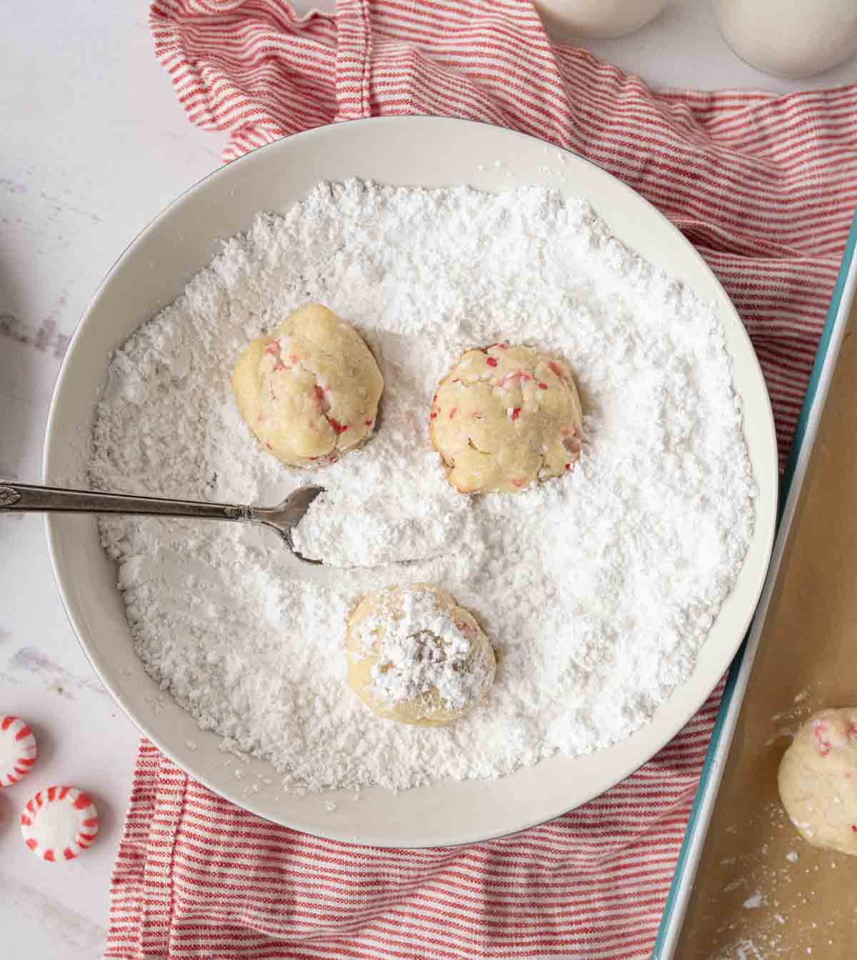A bowl filled with powdered sugar containing three dough balls, one partially coated. A spoon rests in the bowl. The bowl sits on a red-striped cloth next to a baking dish and peppermint candies.