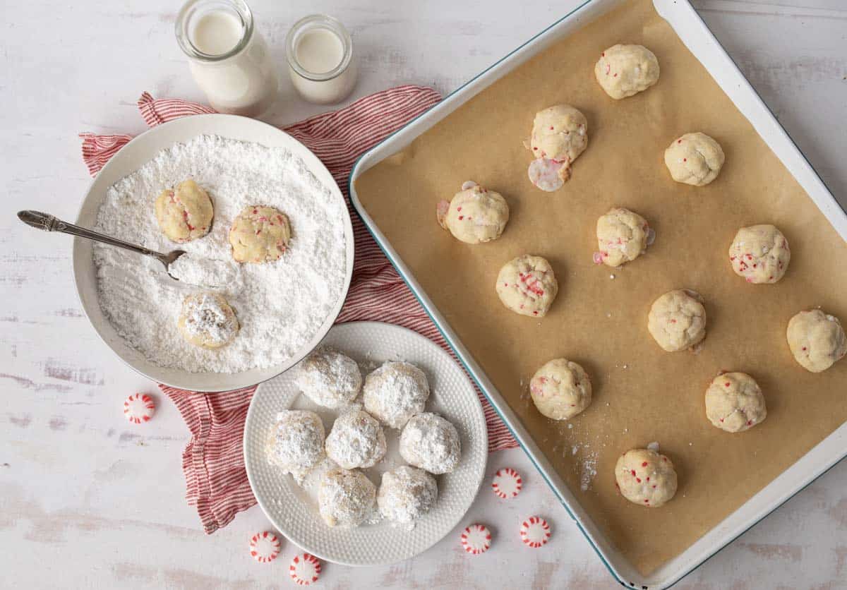Overhead view of unbaked cookie balls on a parchment-lined tray, a bowl with cookie dough being coated in powdered sugar, finished cookies on a plate, two glasses of milk, and peppermint candies on a white surface.