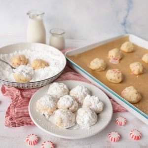 A plate of Peppermint Snowball Cookies dusted with powdered sugar sits on a table beside a bowl of powdered sugar, unbaked cookies, bottles of milk, and peppermint candies, all arranged on a red-striped cloth.