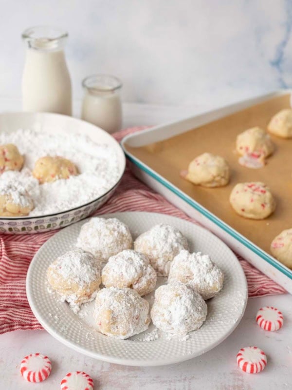 A plate of Peppermint Snowball Cookies dusted with powdered sugar sits on a table beside a bowl of powdered sugar, unbaked cookies, bottles of milk, and peppermint candies, all arranged on a red-striped cloth.