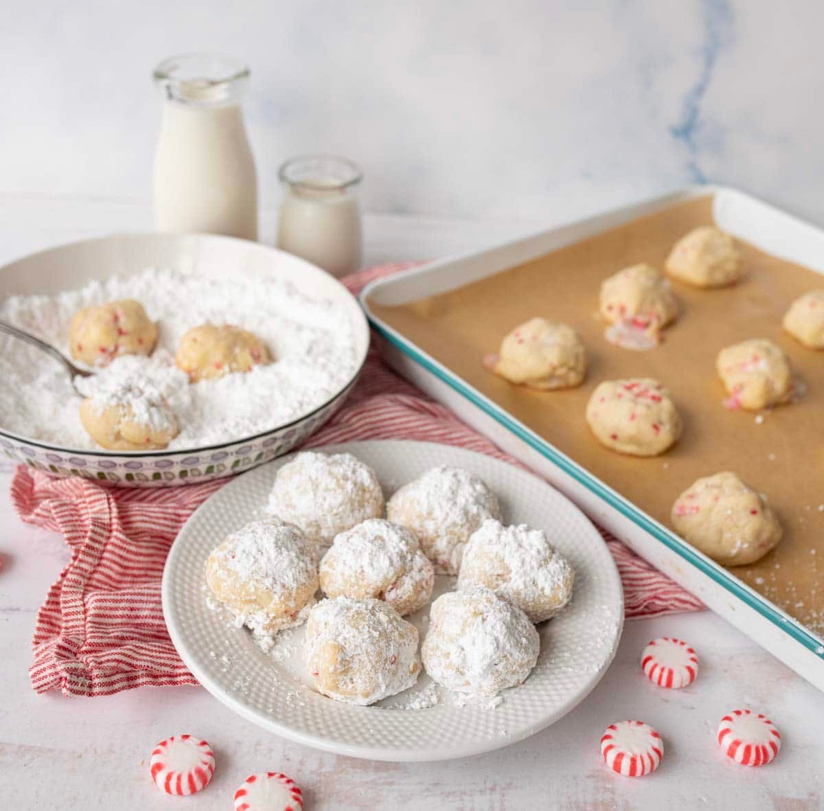 A plate of Peppermint Snowball Cookies dusted with powdered sugar sits on a table beside a bowl of powdered sugar, unbaked cookies, bottles of milk, and peppermint candies, all arranged on a red-striped cloth.