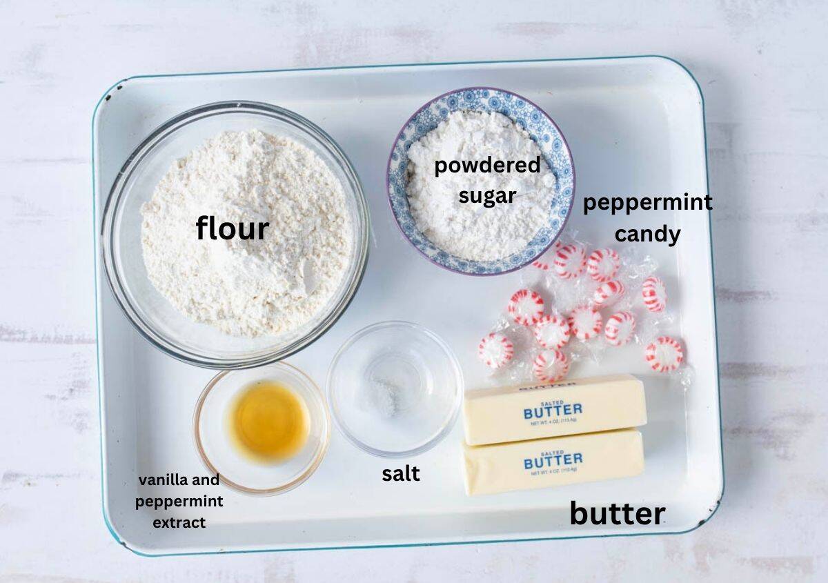 A tray with labeled ingredients for baking: a bowl of flour, a bowl of powdered sugar, peppermint candies, two sticks of butter, vanilla and peppermint extract, and a small dish of salt.