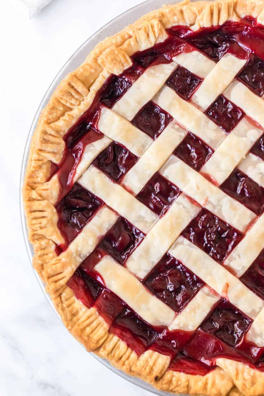 A close-up of a cherry pie with a golden, flaky lattice crust—showcasing expert how to make pie crust techniques—and vibrant cherry filling, displayed on a white marble surface.