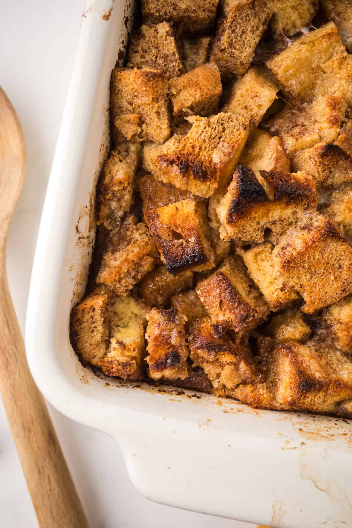A close-up of baked bread pudding in a white dish, showing golden-brown, toasted bread cubes. A wooden spoon is placed next to the dish on a white surface.