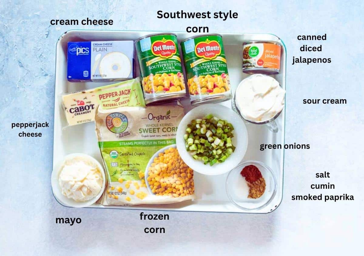 A tray containing ingredients for a dip: canned southwest style corn, canned diced jalapenos, sour cream, sliced green onions, seasonings (salt, cumin, smoked paprika), frozen corn, mayonnaise, pepperjack cheese, and cream cheese.