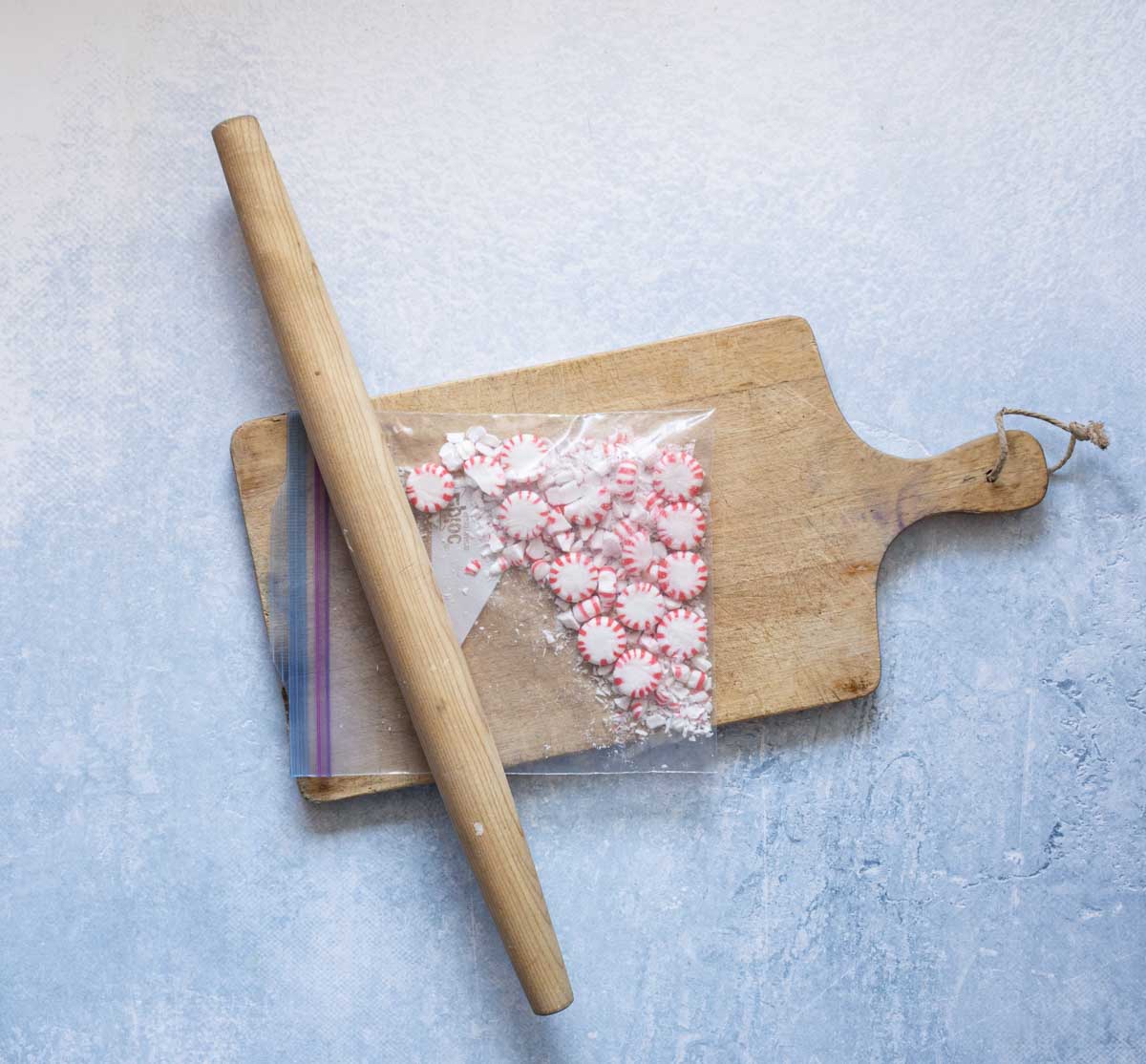 A wooden rolling pin rests on a cutting board with a plastic bag of peppermint candies, some of which are crushed, on a light blue surface.