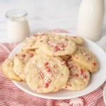 A plate of white chocolate peppermint cookies topped with crushed peppermint candies sits on a red-striped cloth, with a jar and bottle of milk in the background.