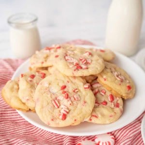 A plate of white chocolate peppermint cookies topped with crushed peppermint candies sits on a red-striped cloth, with a jar and bottle of milk in the background.