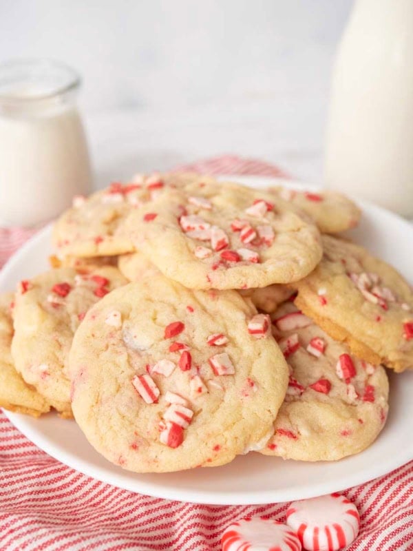 A plate of white chocolate peppermint cookies topped with crushed peppermint candies sits on a red-striped cloth, with a jar and bottle of milk in the background.