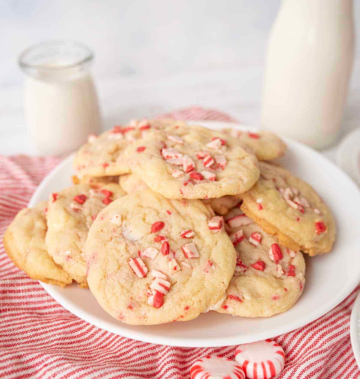A plate of white chocolate peppermint cookies topped with crushed peppermint candies sits on a red-striped cloth, with a jar and bottle of milk in the background.
