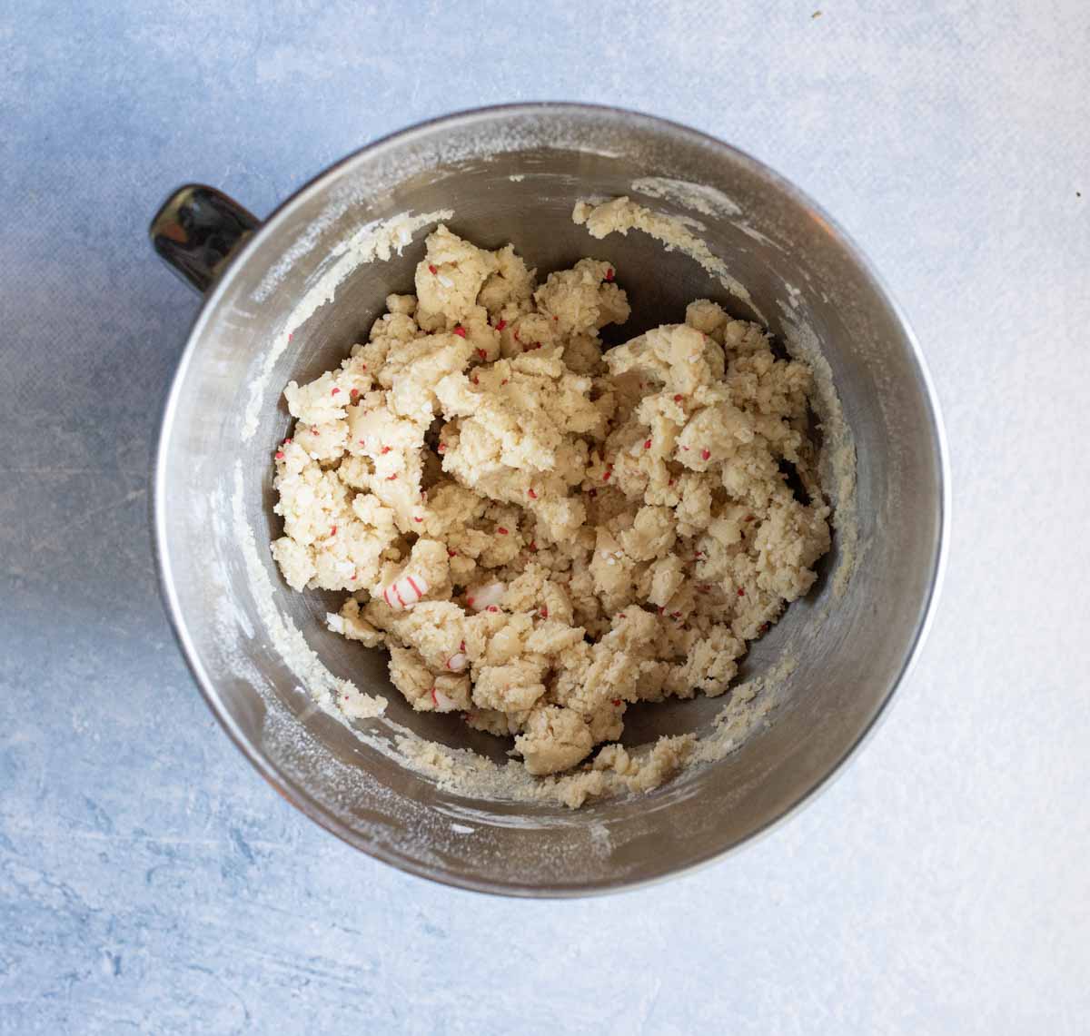 A metal mixing bowl filled with chunky cookie dough, containing small red and white bits, sits on a light blue textured surface.