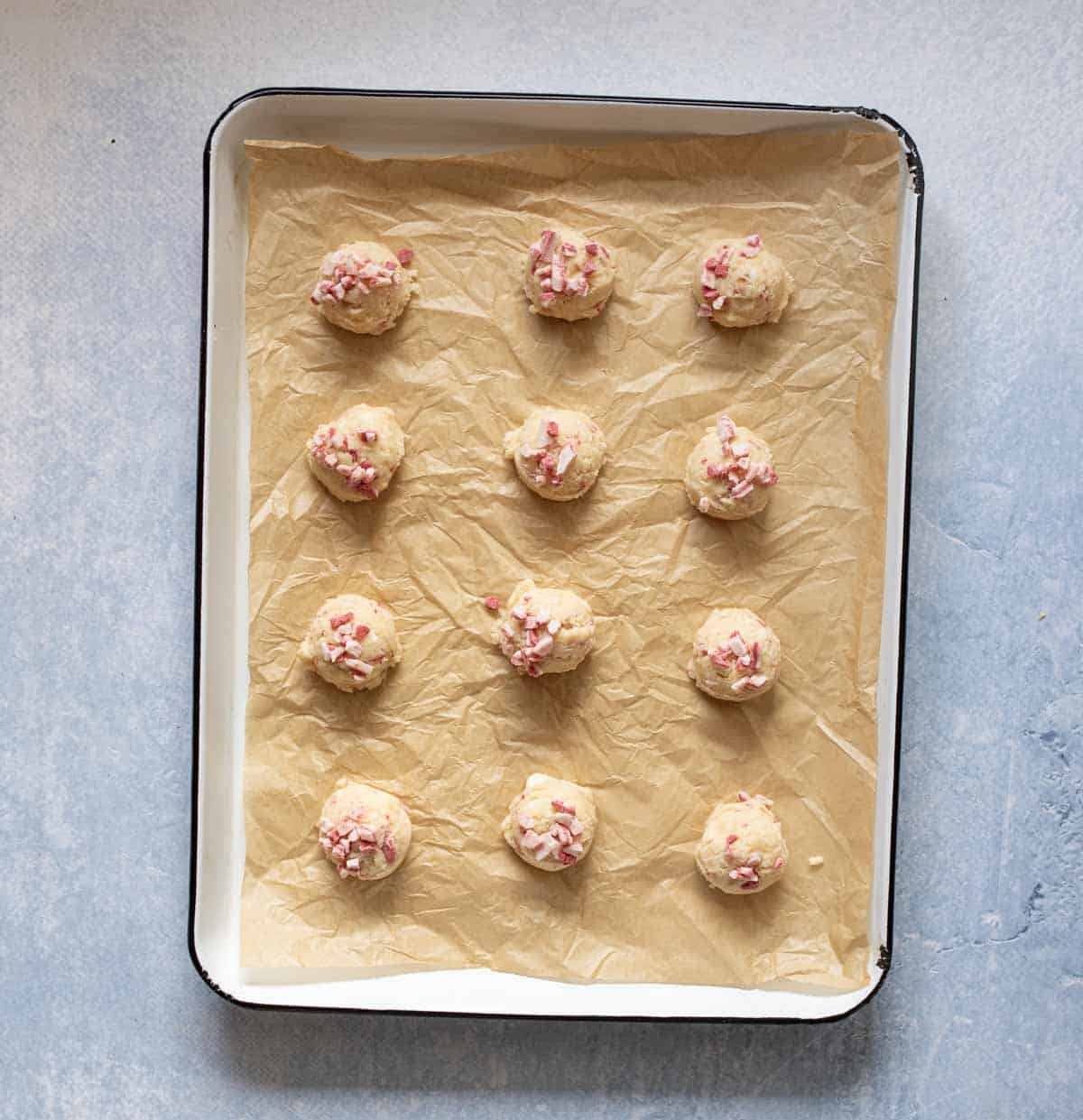Twelve unbaked cookie dough balls with pink and white pieces on top are arranged on a baking tray lined with brown parchment paper. The tray is set on a light blue surface.