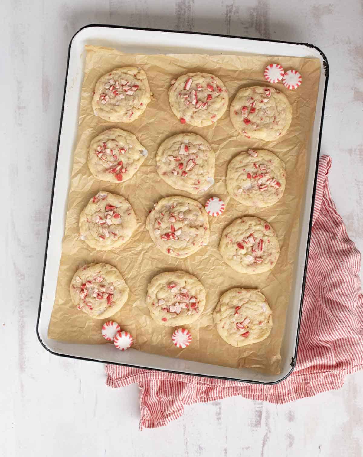 A baking tray lined with parchment paper holds twelve cookies topped with crushed peppermint candies. A few whole peppermint candies are scattered on the tray, and a red-striped cloth is nearby on a white surface.