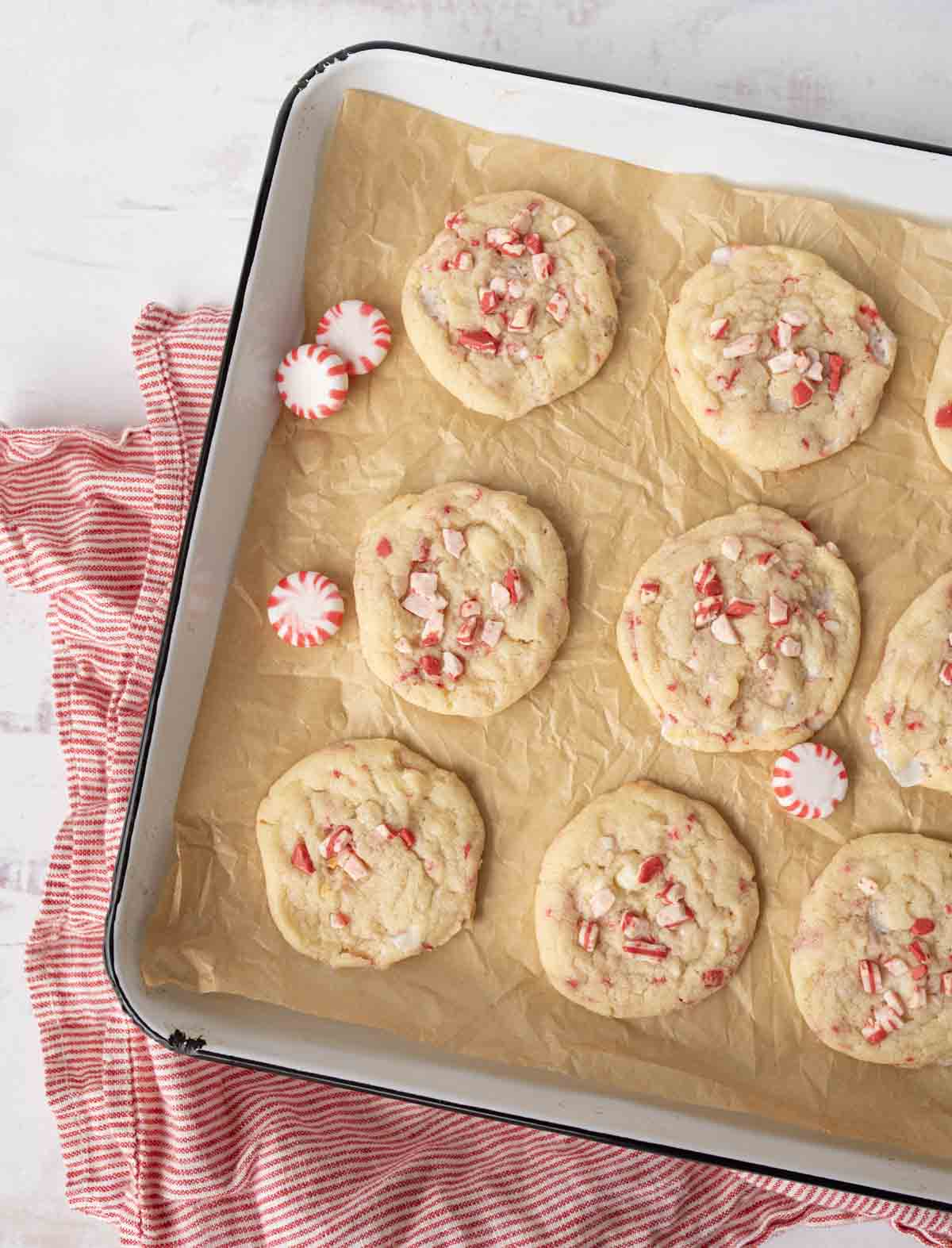 A baking tray lined with parchment paper holds eight cookies topped with crushed peppermint candies, with whole peppermint candies nearby. The tray rests on a red and white striped cloth.