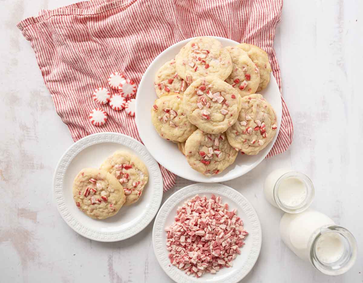 A plate of peppermint cookies sits on a red-striped cloth, with small bowls of crushed peppermint and whole peppermint candies nearby, and two bottles of milk on a white surface.
