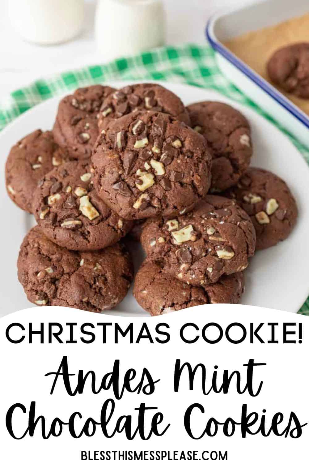 A plate of andes mint chocolate cookies sits on a white surface, topped with chopped mint chocolate pieces. A green checkered cloth and a baking tray of fresh andes mint chocolate cookies are in the background.