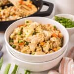 A white bowl filled with chicken fried rice sits in front, showcasing chunks of chicken, peas, carrots, and scrambled eggs—the perfect result after making chicken fried rice. In the background are a skillet with more rice and a bowl of chopped green onions.