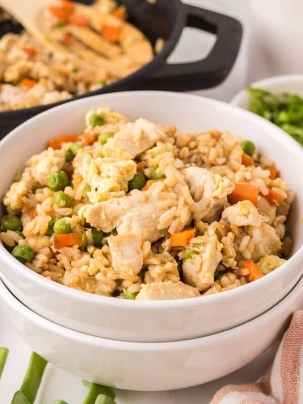 A white bowl filled with chicken fried rice sits in front, showcasing chunks of chicken, peas, carrots, and scrambled eggs—the perfect result after making chicken fried rice. In the background are a skillet with more rice and a bowl of chopped green onions.