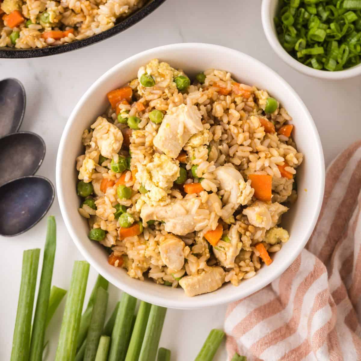 A bowl of chicken fried rice with vegetables, including peas and carrots, sits on a white surface—perfect for making chicken fried rice—near green onions, a striped towel, two spoons, and a small bowl of chopped green onions.