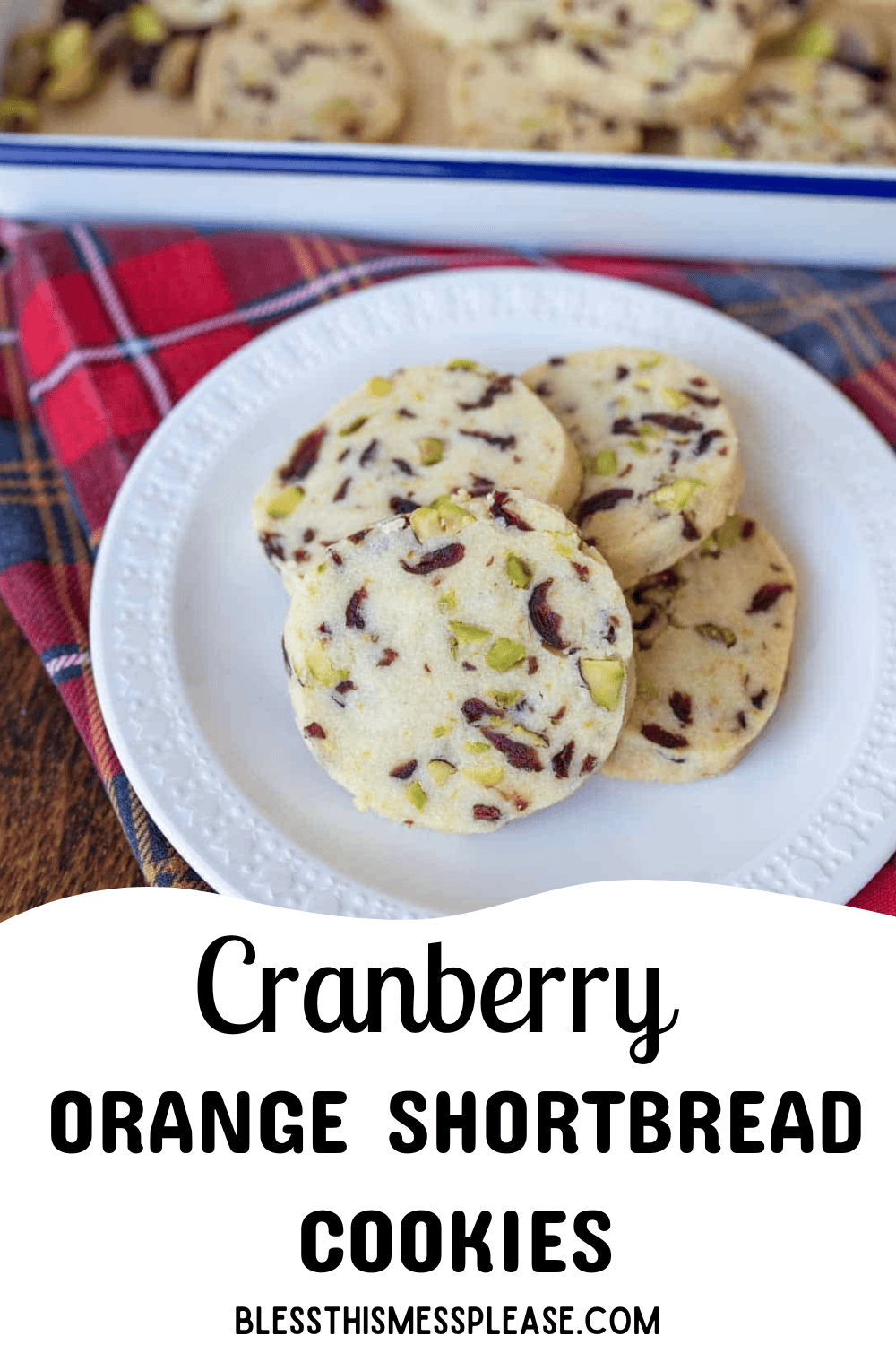 A white plate holds three round cranberry orange shortbread cookies, studded with cranberries and pistachios. More cranberry orange shortbread cookies rest on a baking tray atop a red plaid cloth. Text reads Cranberry Orange Shortbread Cookies.