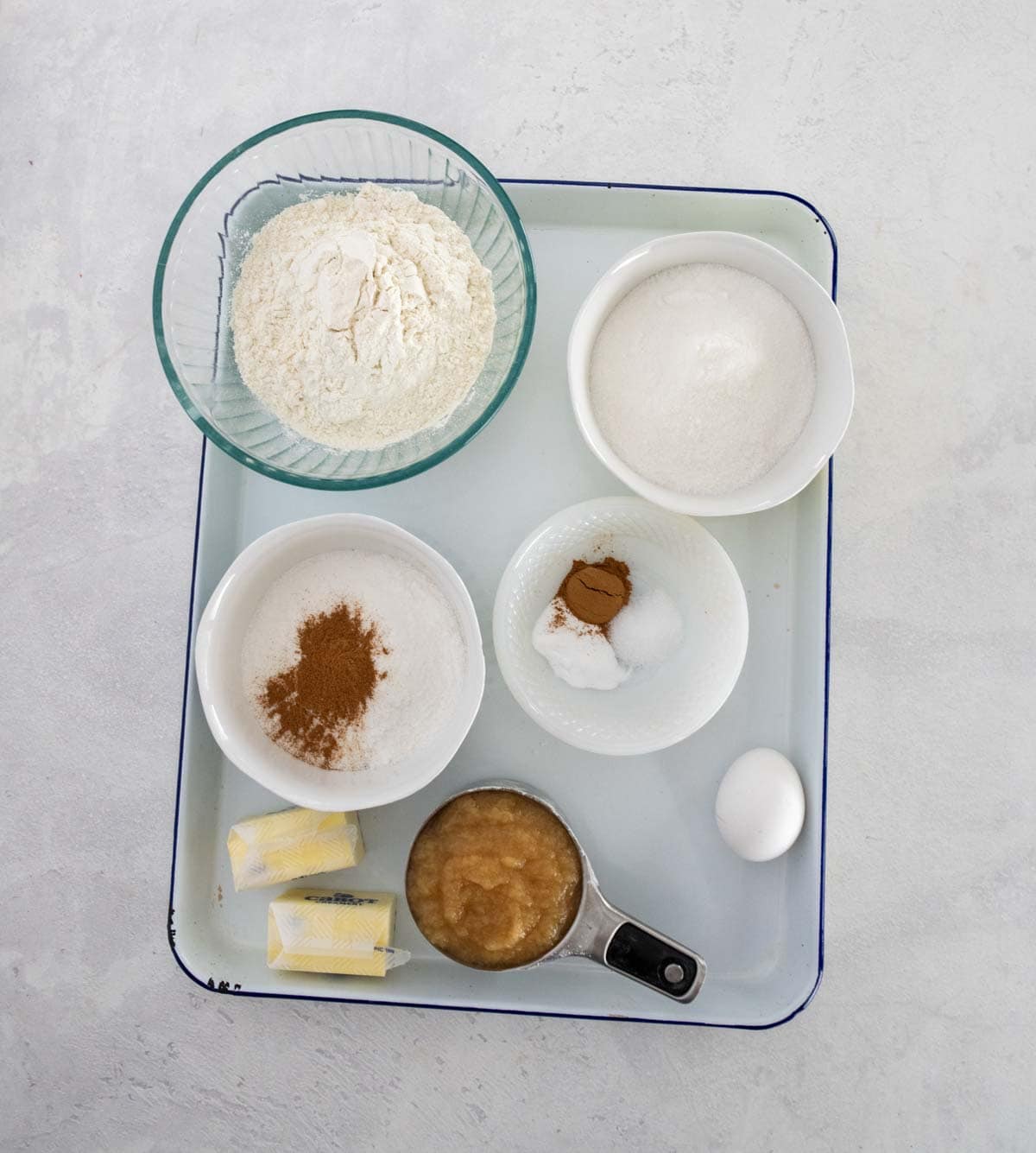 A tray with small bowls containing flour, sugar, cinnamon, baking soda, salt, and applesauce&mdash;perfect for baking an applesauce coffee cake&mdash;plus two butter sticks and a whole egg arranged neatly on a white surface.